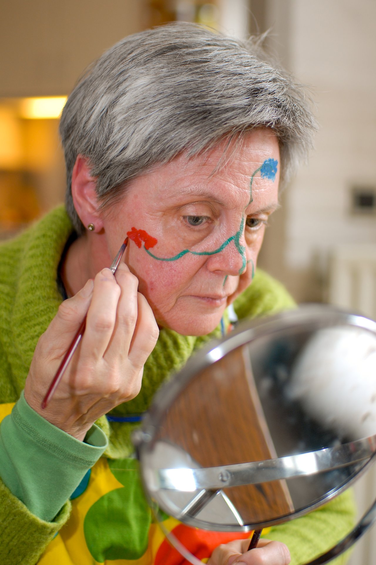 An older woman paints her face with colorful designs while looking into a mirror, preparing for Carnaval.