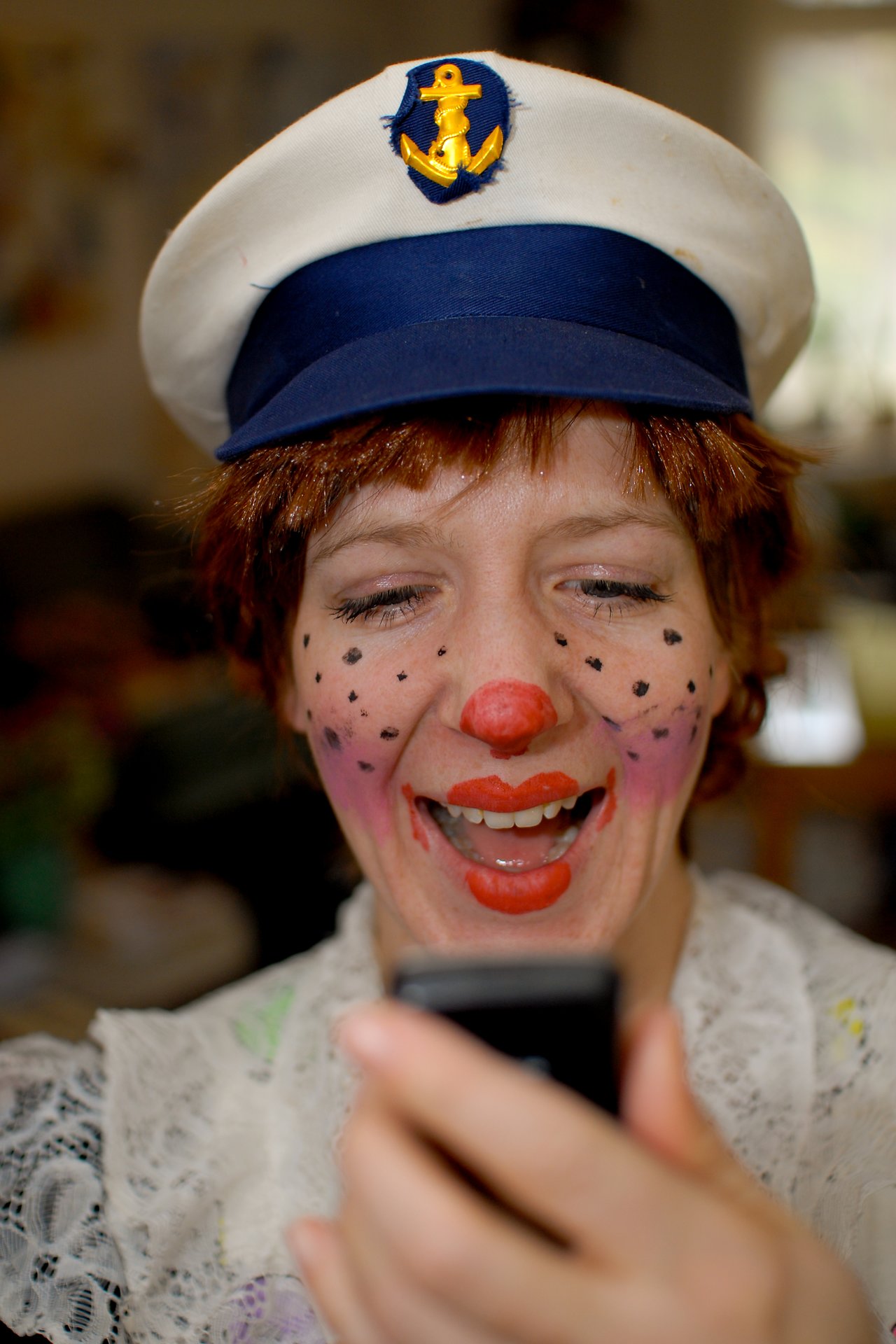 Person in clown makeup and a sailor hat smiles while looking at a phone during a carnival celebration.