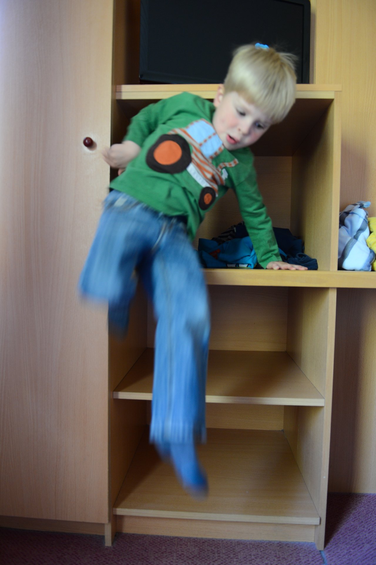 A young child in a green shirt jumps down from a wooden shelf, holding onto the edge for support.