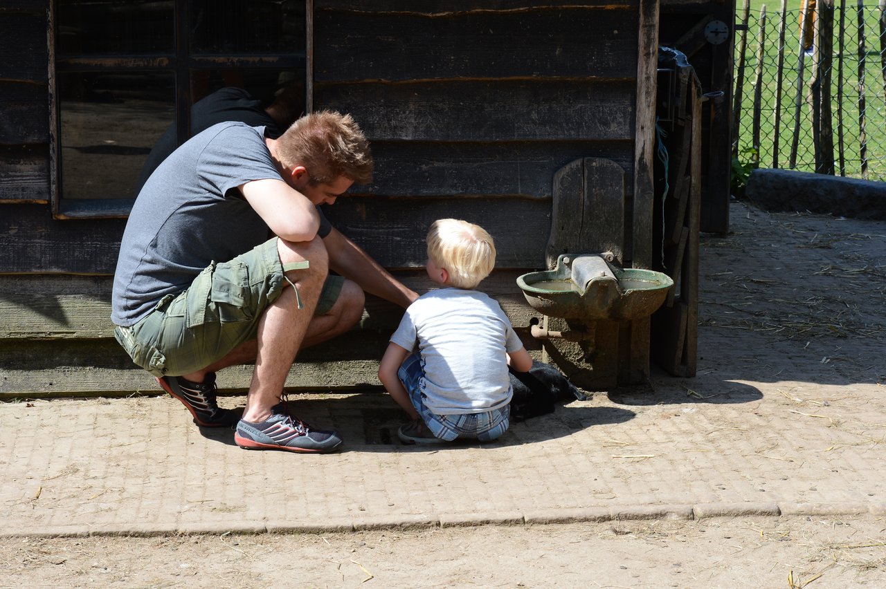 A man and a young child crouch near a wooden structure, looking at something on the ground together.