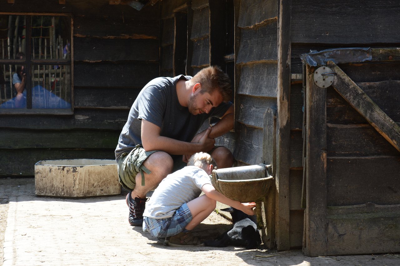 A man and a child crouch near a wooden structure, with the child reaching out to touch a small black animal.