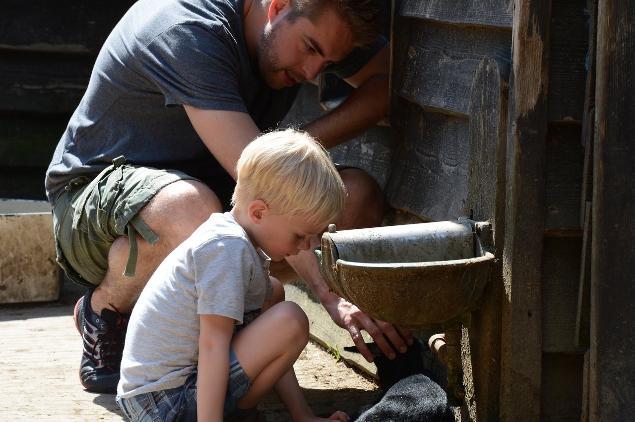 A man and a young boy crouch near a water trough, petting a small black animal.
