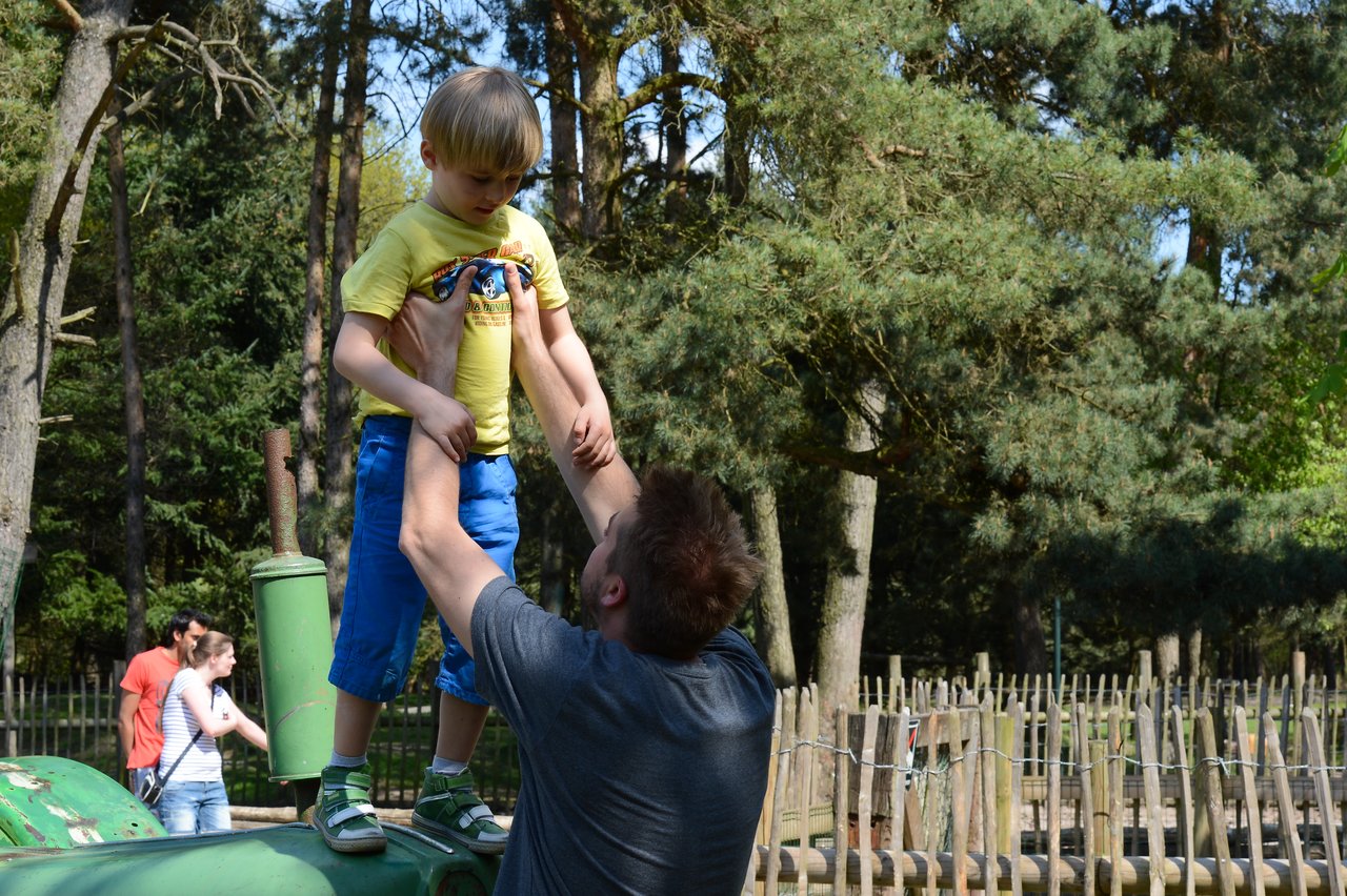 An adult lifts a child down from a green tractor in an outdoor park with trees and wooden fences.