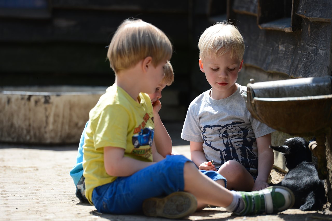 Three young children sit on the ground, interacting with a small black goat near a water trough.