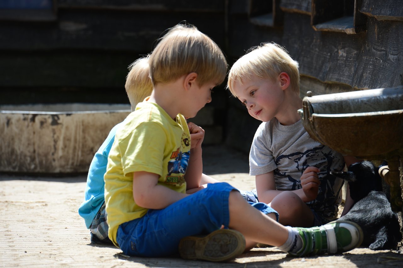 Three young children sit on the ground, interacting with a small black goat near a water trough.