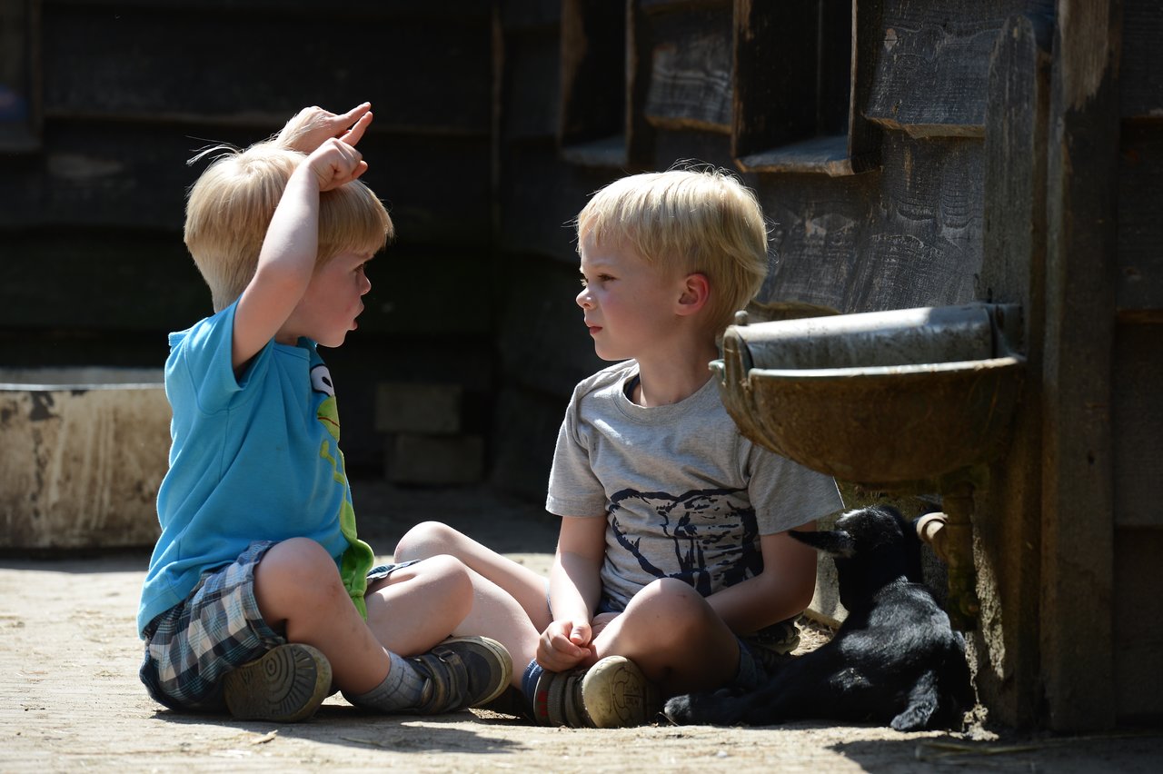 Two young children sit on the ground, talking, while a small black goat rests nearby near a water trough.