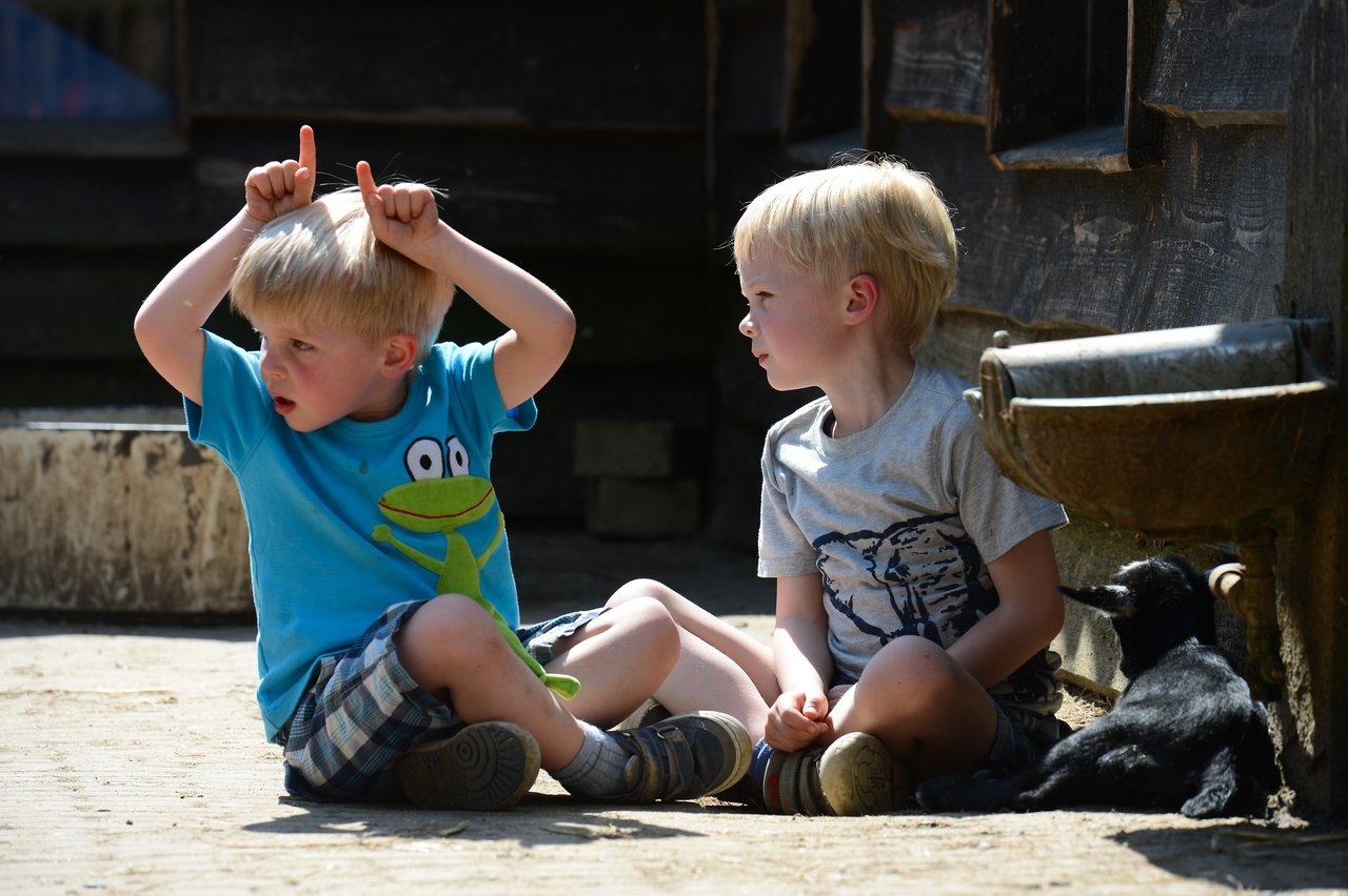 Two young boys sit on the ground, one making playful hand gestures, while a small black goat rests nearby.