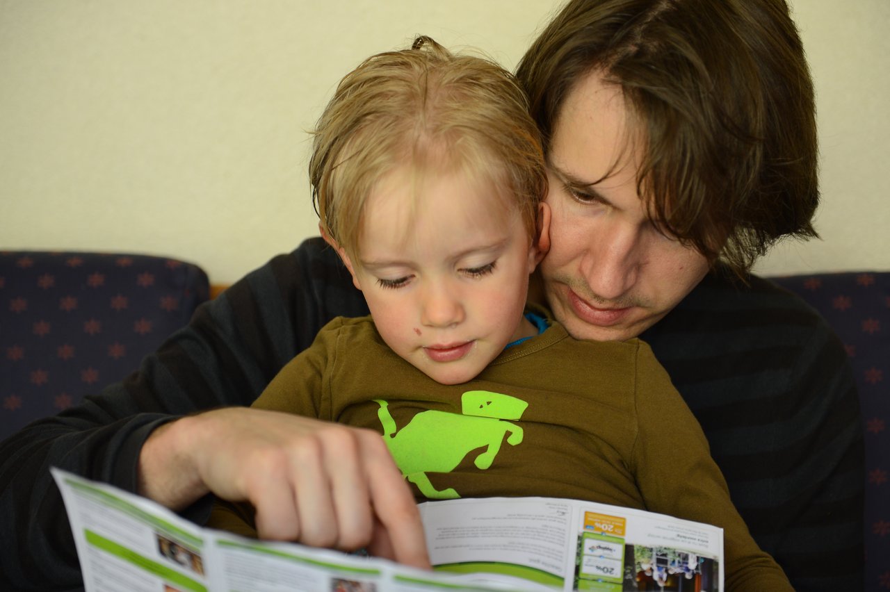 An adult and a child sit closely together, looking at a brochure while the adult points at something.