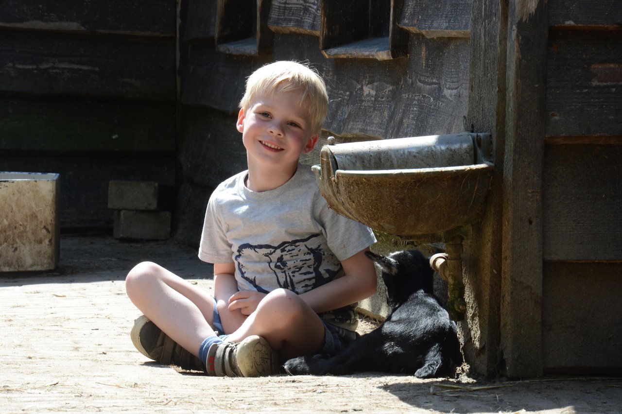 A young child sits on the ground, smiling, next to a small black goat drinking from a water trough.