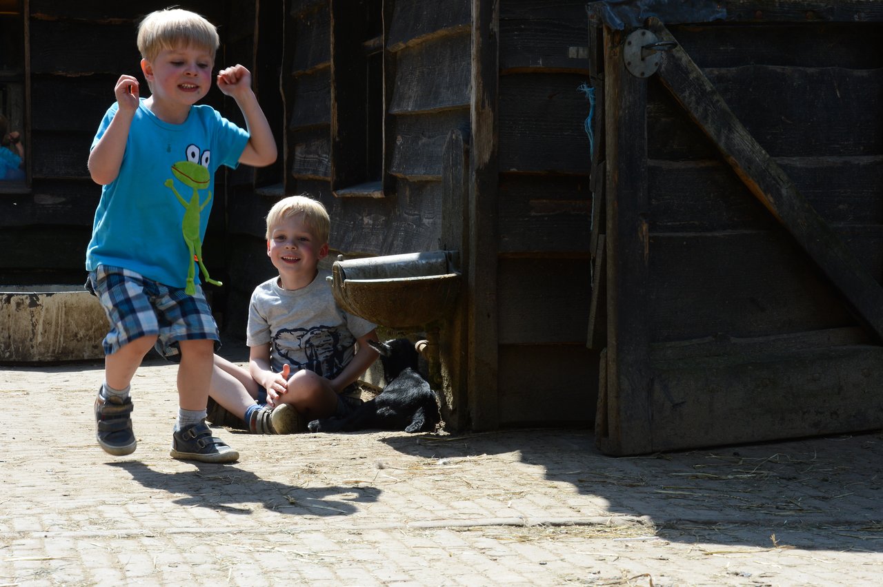 A young boy jumps with excitement while another boy sits on the ground, smiling near a wooden barn.