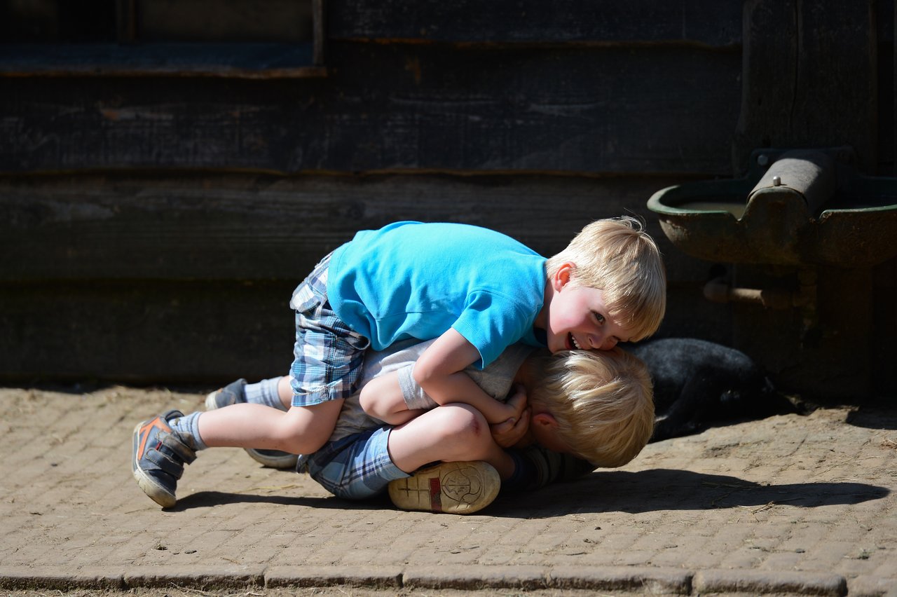Two young boys in matching outfits playfully wrestle on the ground, smiling and laughing in the sunlight.