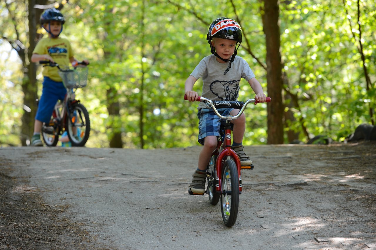 Two children ride bicycles on a dirt path in a wooded area, both wearing helmets for safety.
