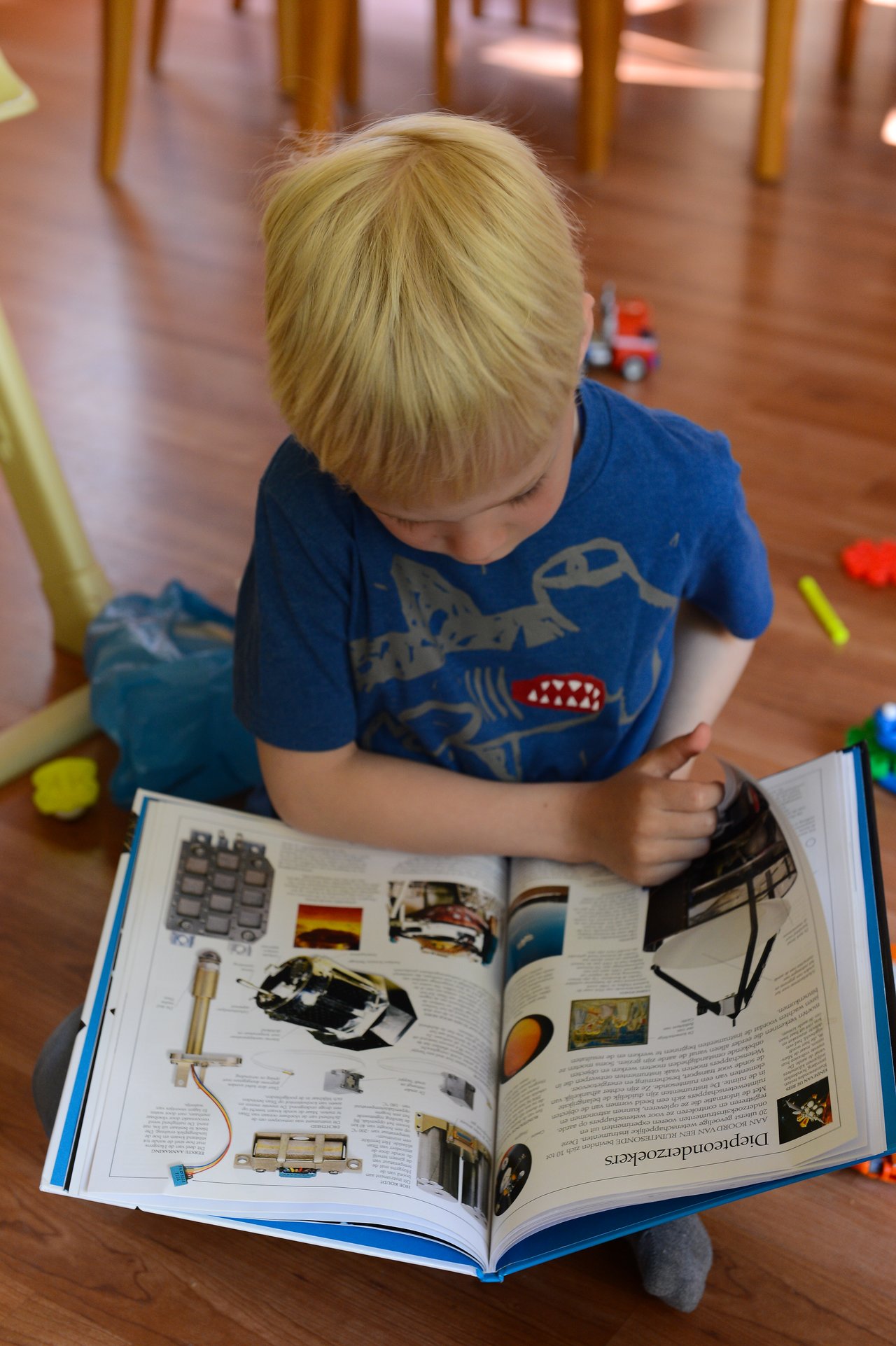A young child in a blue shirt sits on the floor, reading a large book about space and technology.