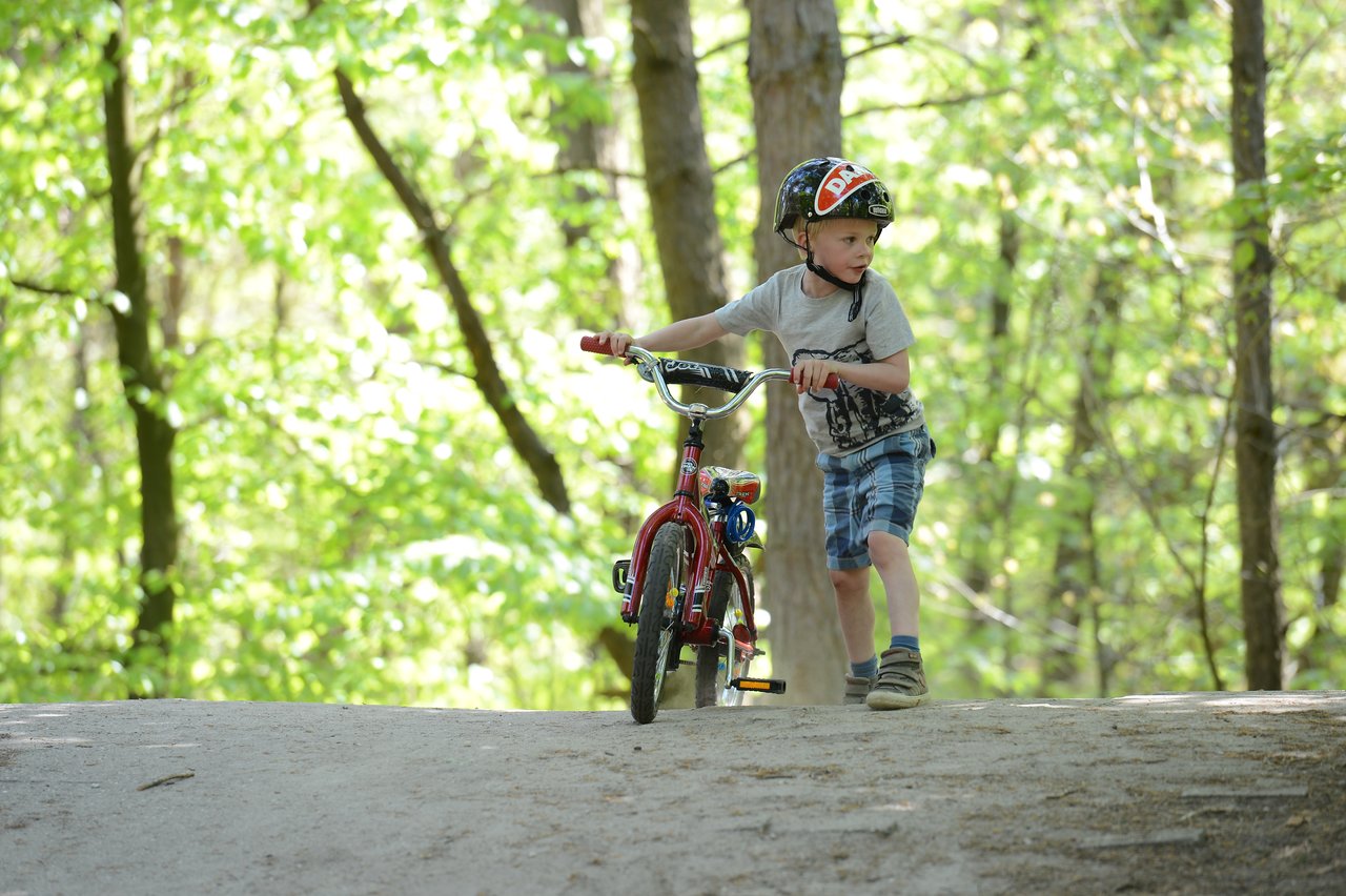 A child wearing a helmet walks a red bicycle up a dirt path in a wooded area.