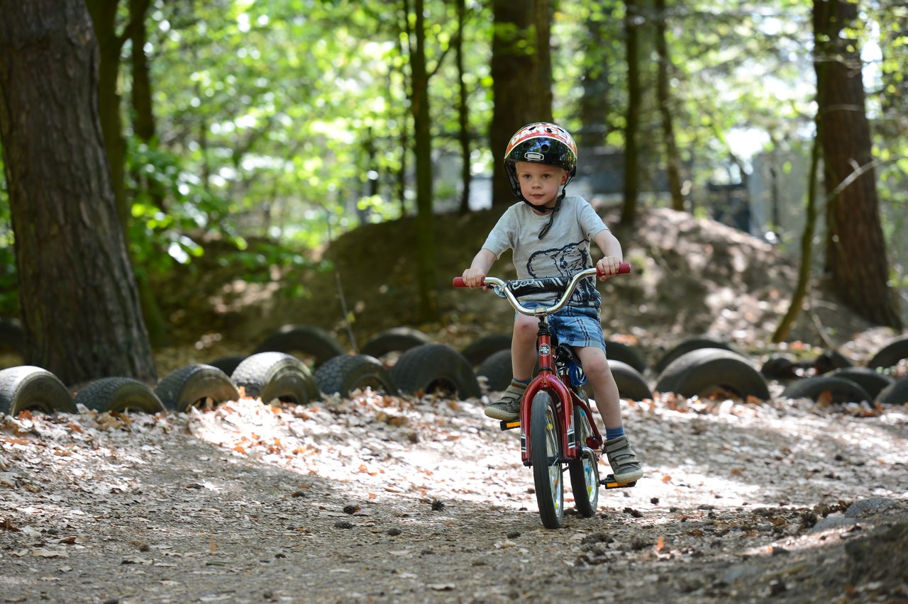 A young child wearing a helmet rides a red bicycle on a dirt path surrounded by trees and tires.
