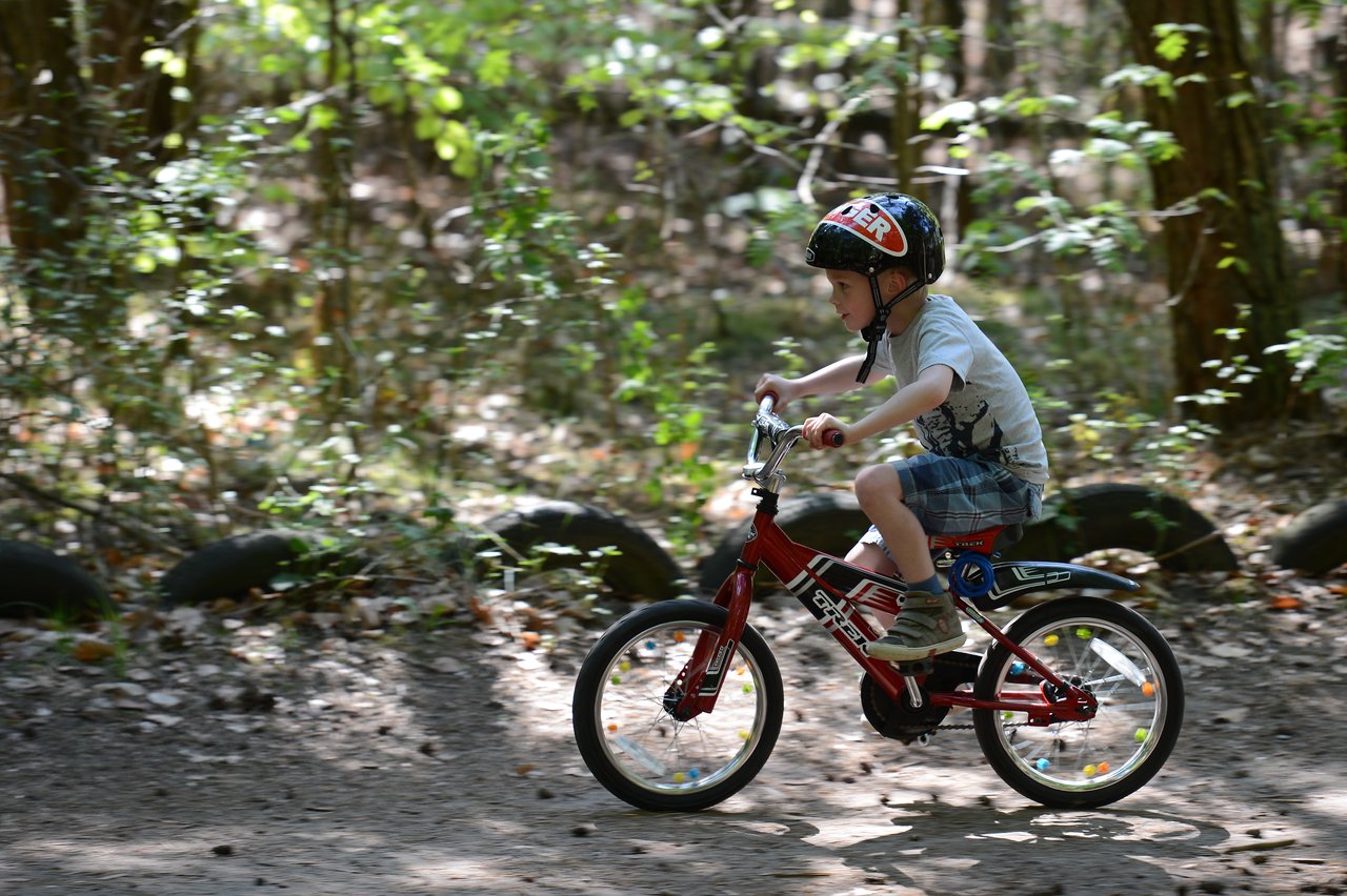 A young child wearing a helmet rides a red bicycle on a dirt path in a wooded area.
