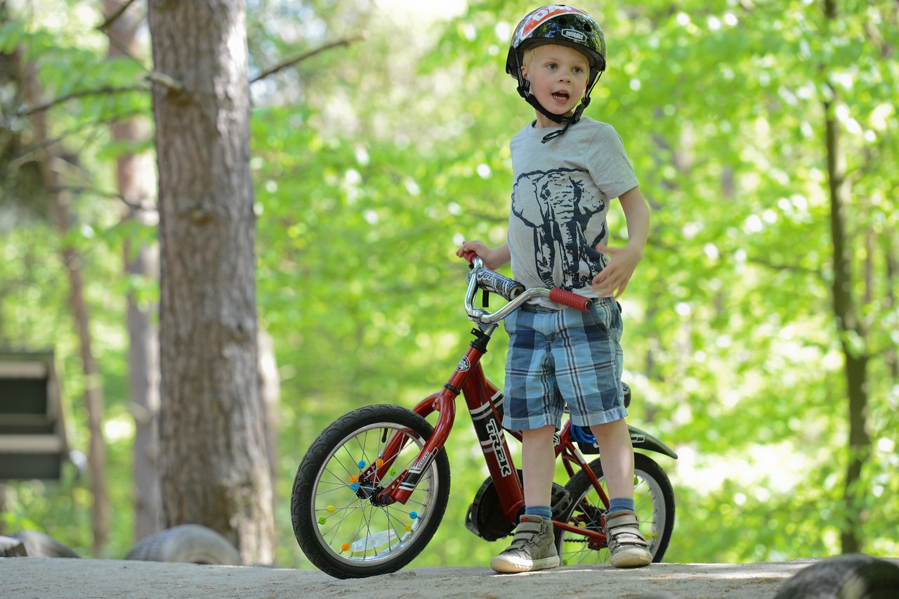 A young child wearing a helmet stands with a red bicycle on a dirt path in a wooded area.