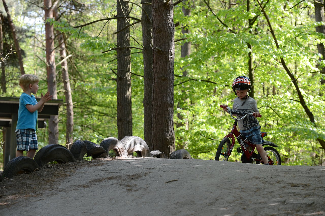 A child wearing a helmet rides a bicycle down a dirt path while another child claps nearby.