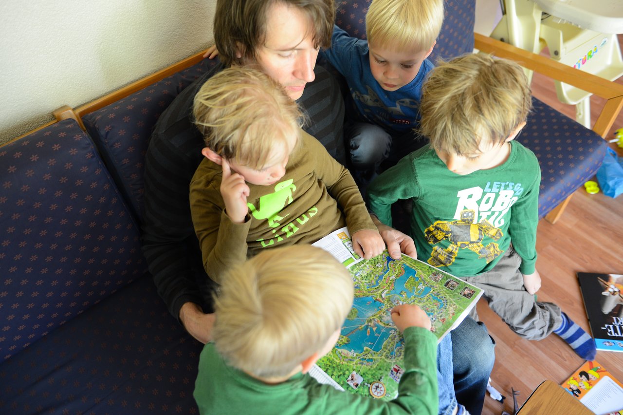 An adult and four children sit on a couch, looking at and pointing to a detailed map together.