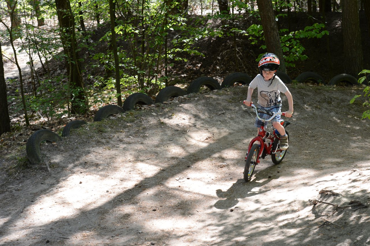 A child wearing a helmet rides a red bicycle on a dirt path surrounded by trees and tires.