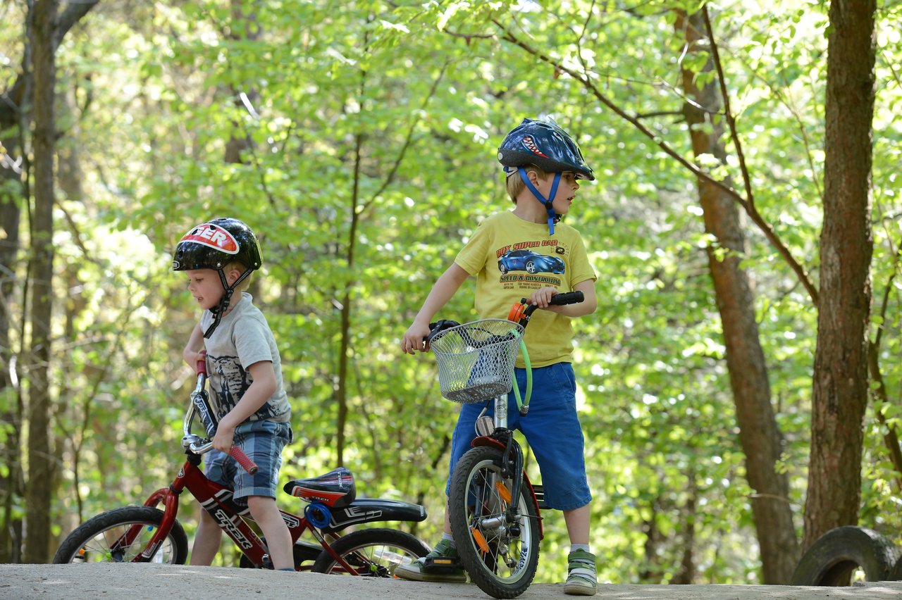Two children wearing helmets ride bicycles on a dirt path in a wooded area, looking in different directions.
