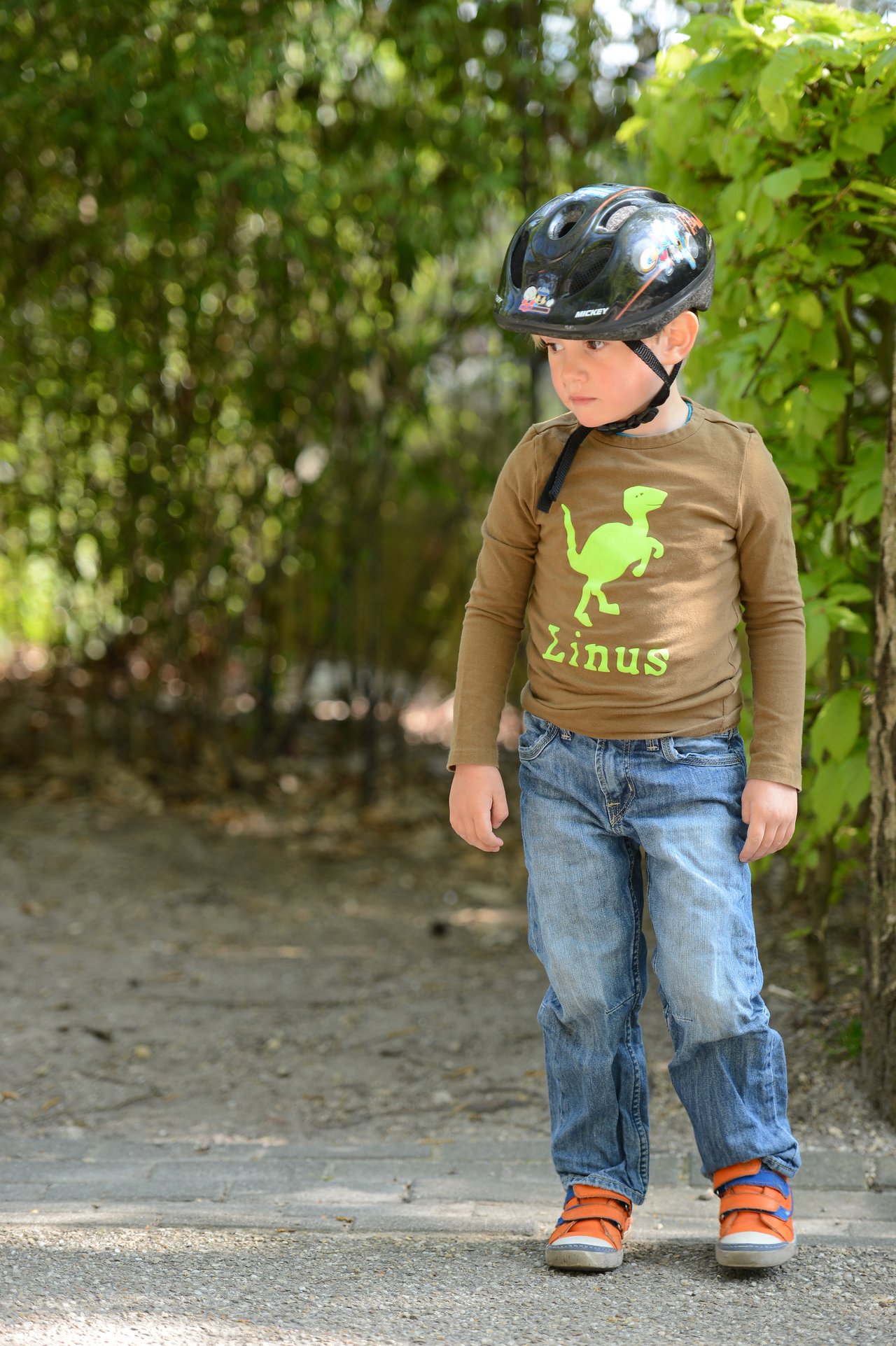A young child wearing a bike helmet and casual clothes stands on a path, looking to the side.