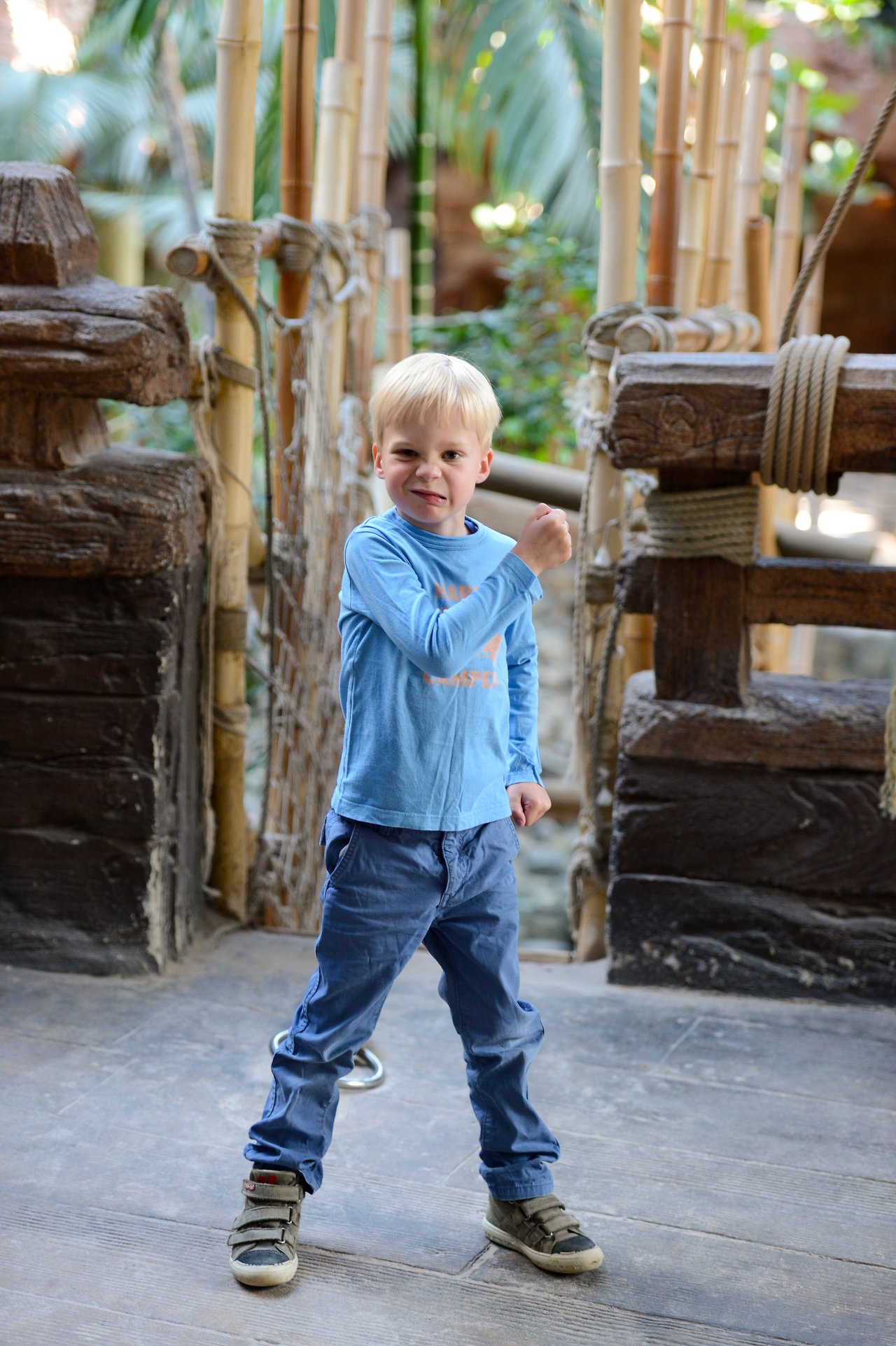 A young boy in a blue shirt stands on a wooden bridge, raising his arm with a playful expression.