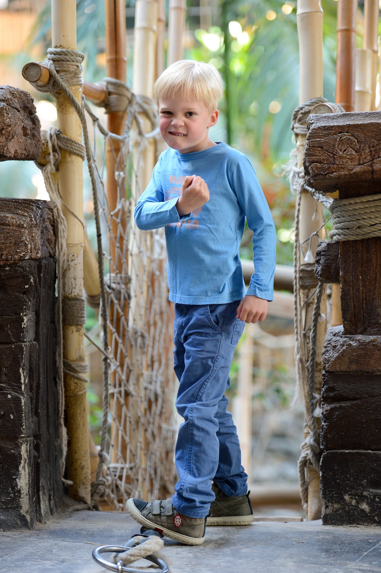 A young child in a blue shirt stands on a wooden bridge, smiling and making a playful fist gesture.