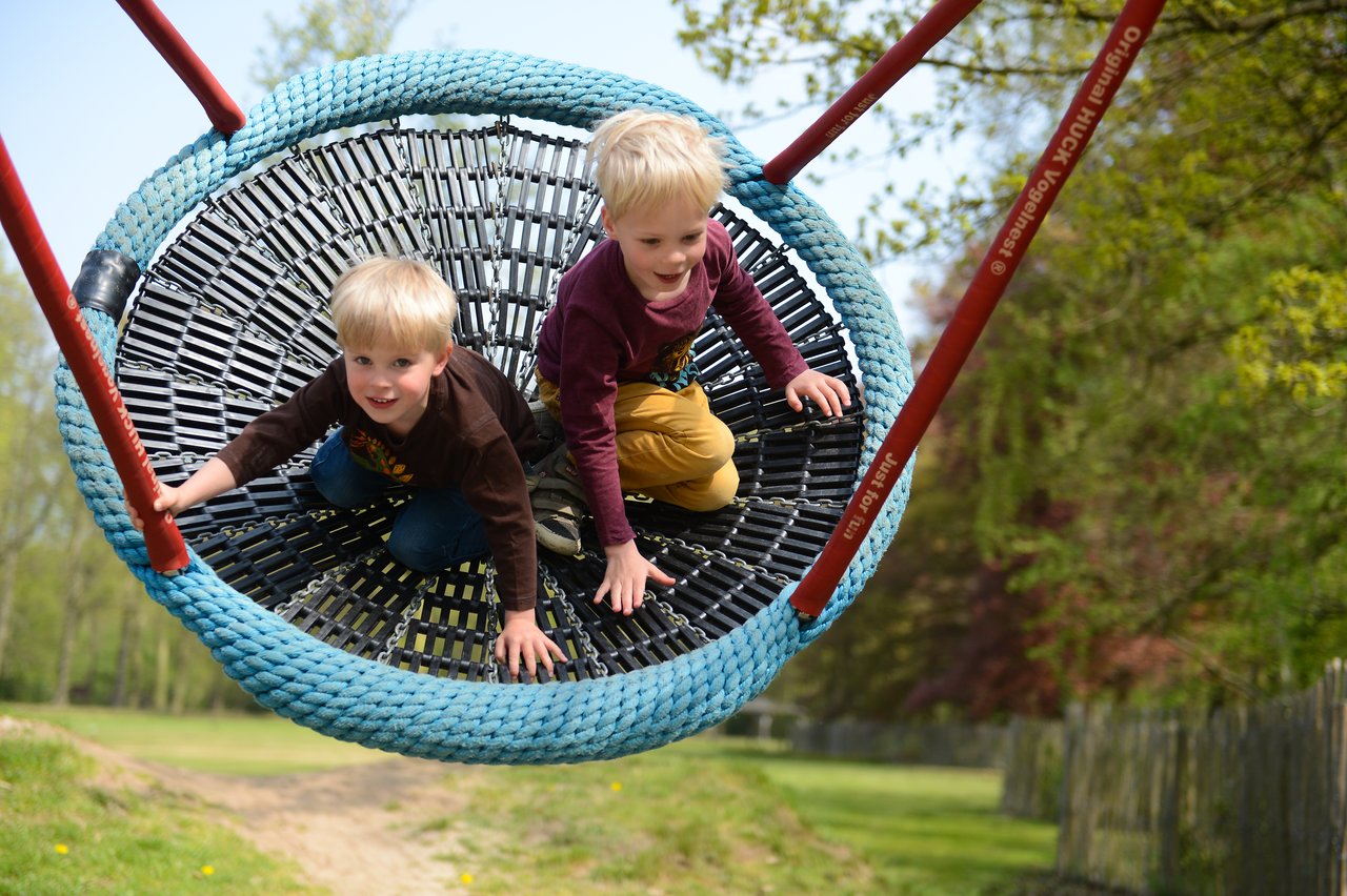 Two young children play on a large rope swing, holding on and smiling as they swing forward.