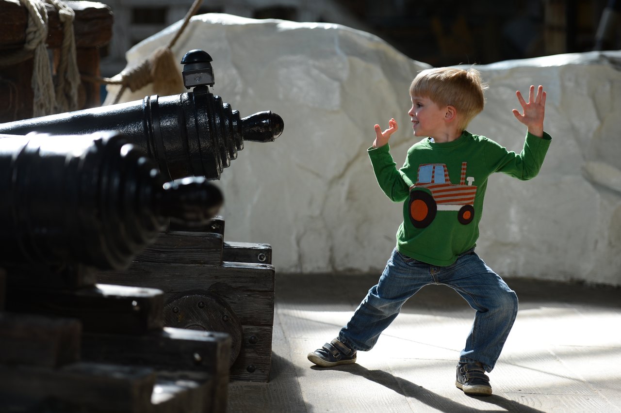 A young child in a green shirt playfully poses in front of two large black cannons indoors.