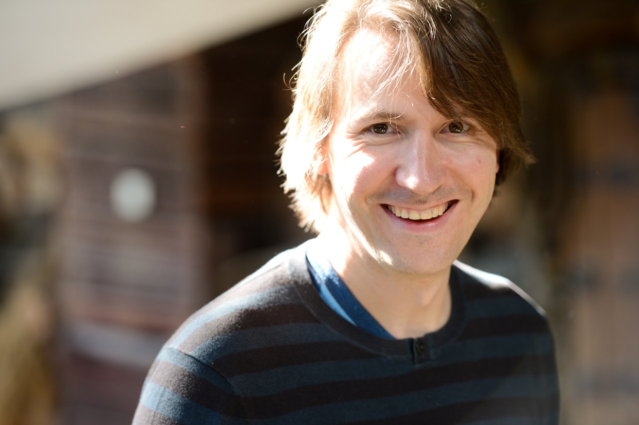 A person with medium-length hair and a striped sweater smiles at the camera in natural lighting.
