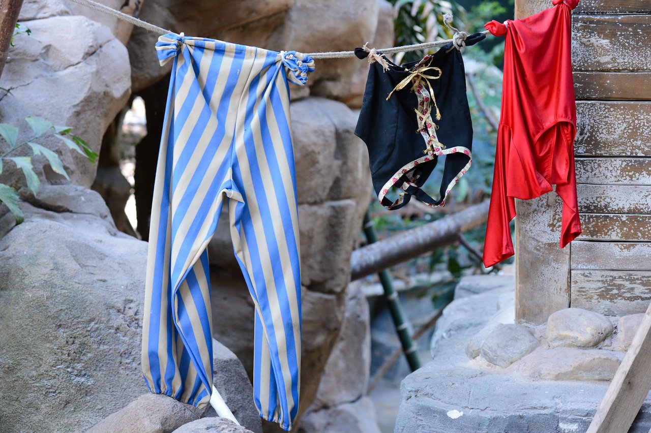 Striped pants, black shorts, and a red garment hang on a clothesline outdoors near rocks and wooden structures.
