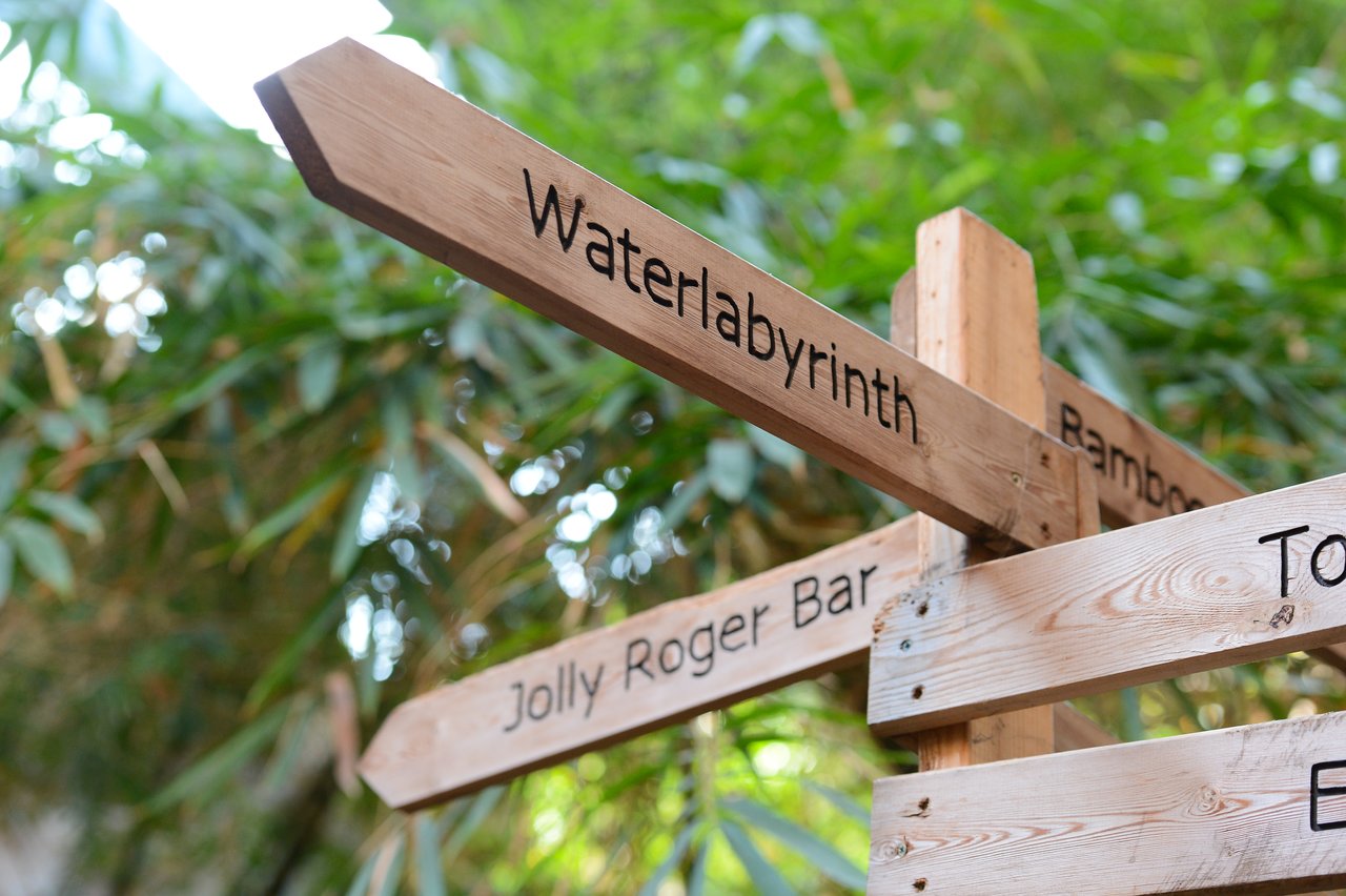 Wooden signpost with multiple directional signs, including "Waterlabyrinth" and "Jolly Roger Bar", surrounded by green foliage.