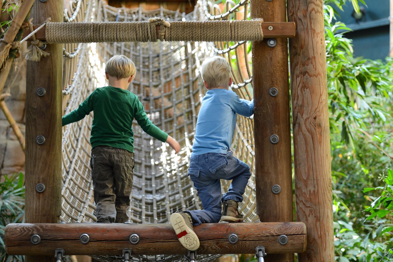 Two children climb a rope bridge at an outdoor play area, holding onto the netting for support.