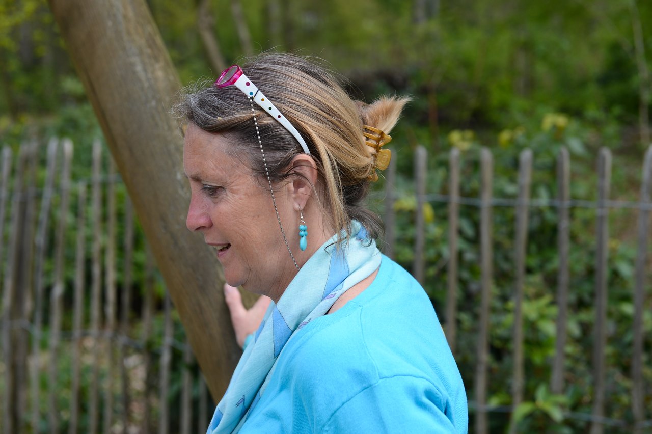 A woman in a blue shirt and scarf stands outdoors, looking to the side with sunglasses on her head.