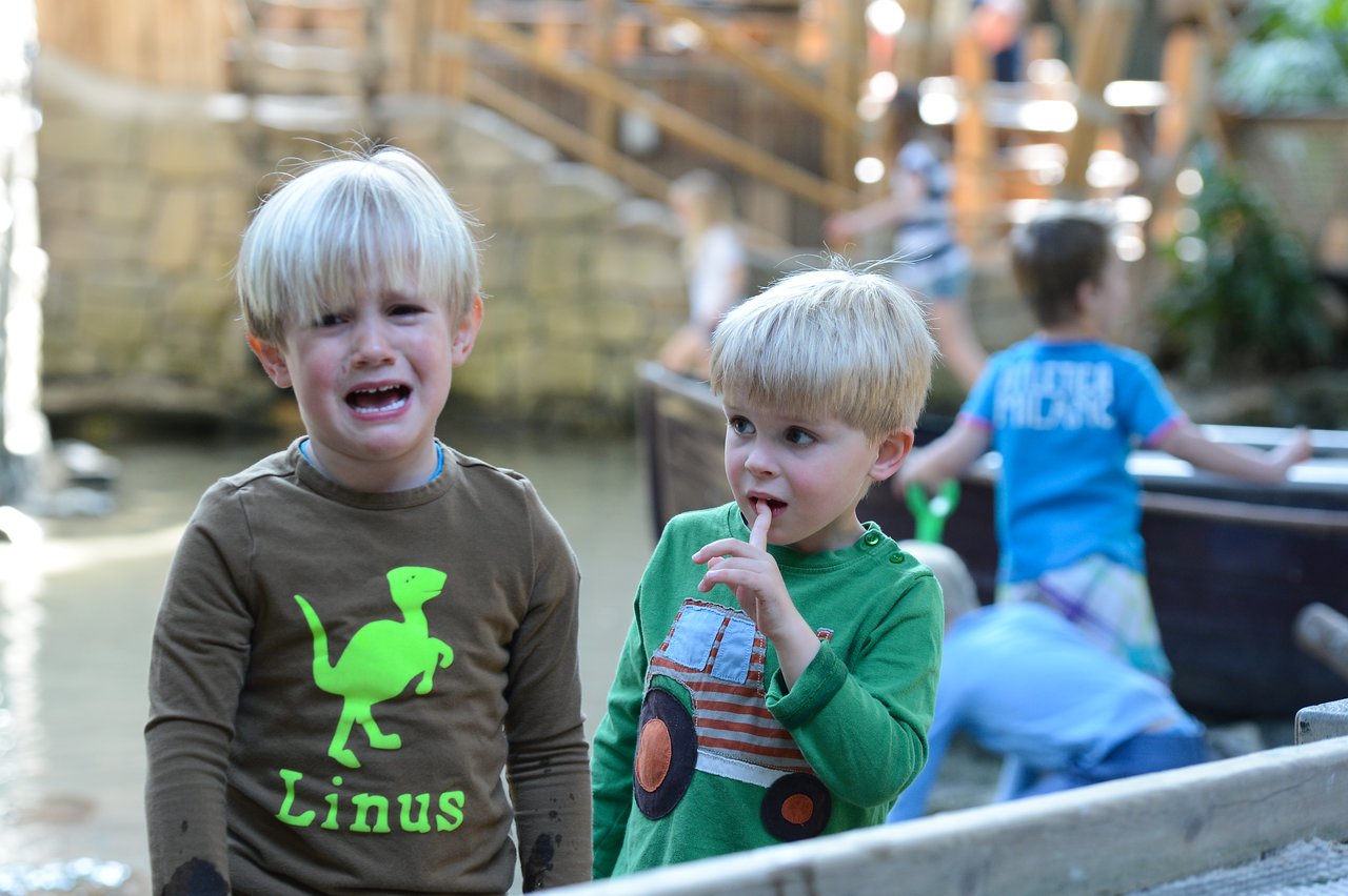 A young boy in a brown shirt is crying, while another boy in green watches with his finger on his lips.