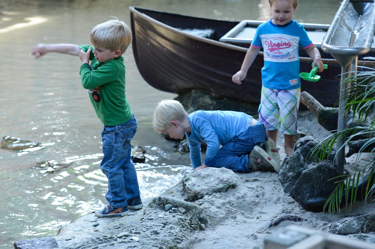 Three young children play near the water; one throws something, another crouches, and the third holds a toy shovel.