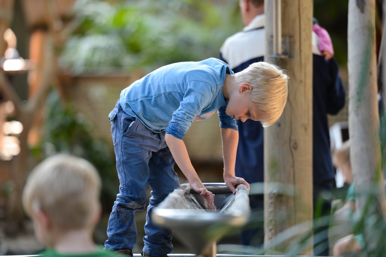 A young boy in a blue shirt leans over a water play structure, interacting with the flowing water.
