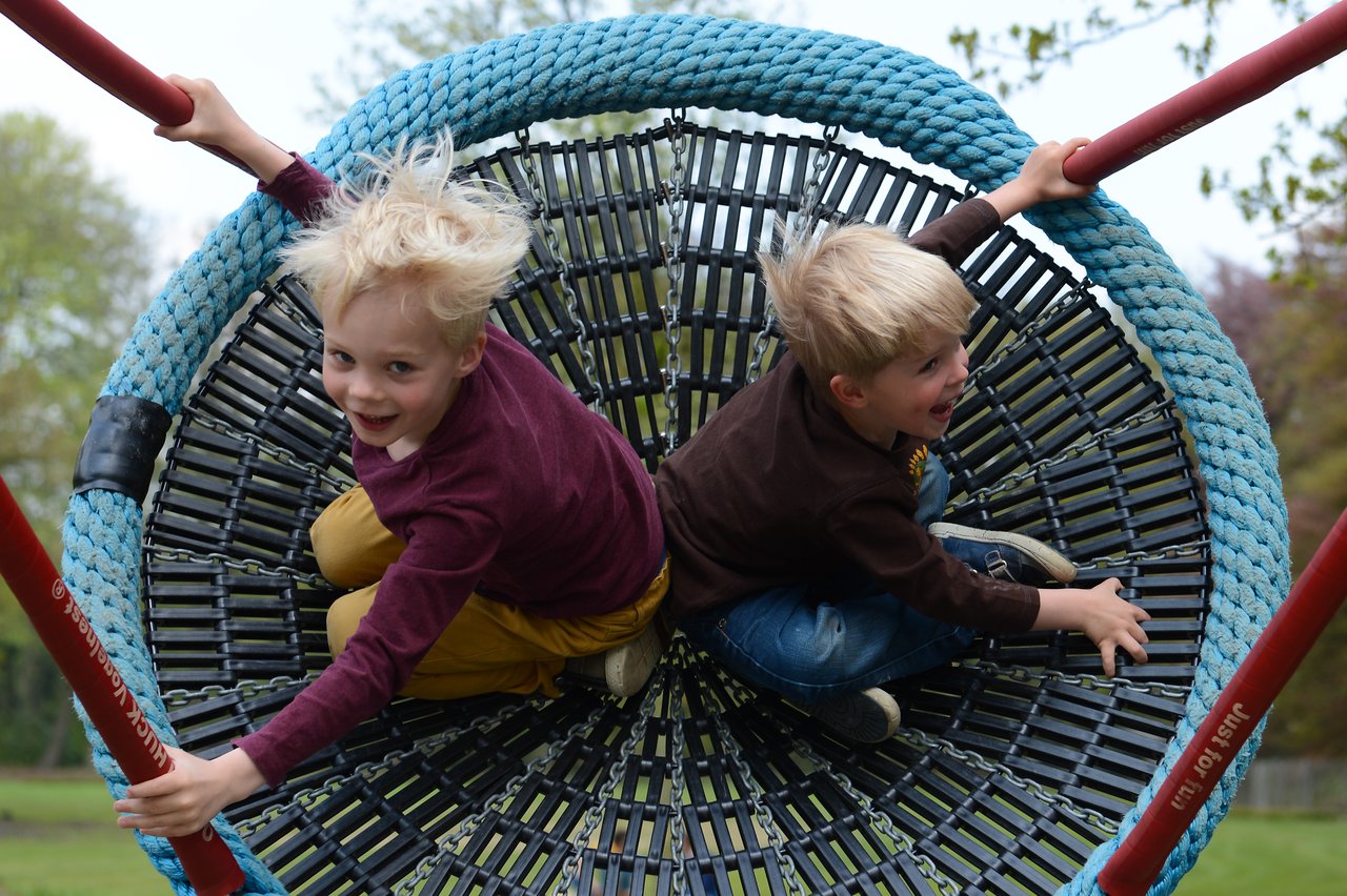 Two children smile and hold onto a large rope swing while playing at a park.
