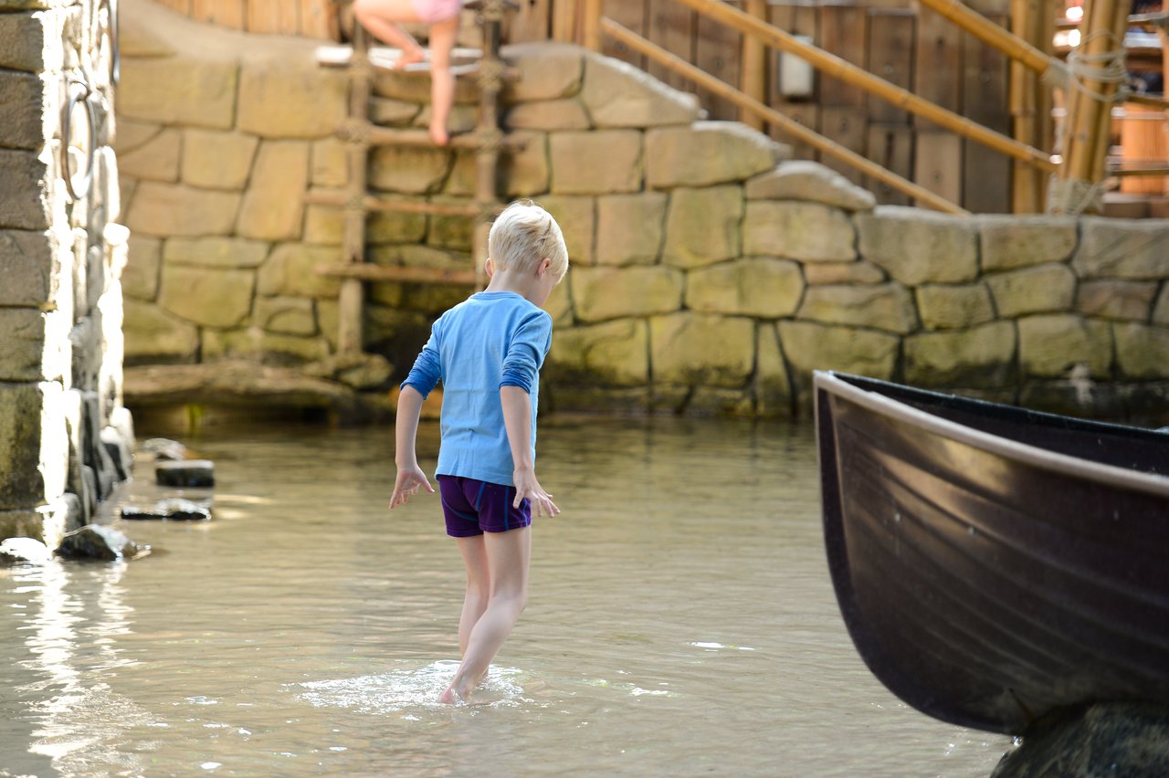 A child in a blue shirt and purple shorts walks through shallow water near a black boat.