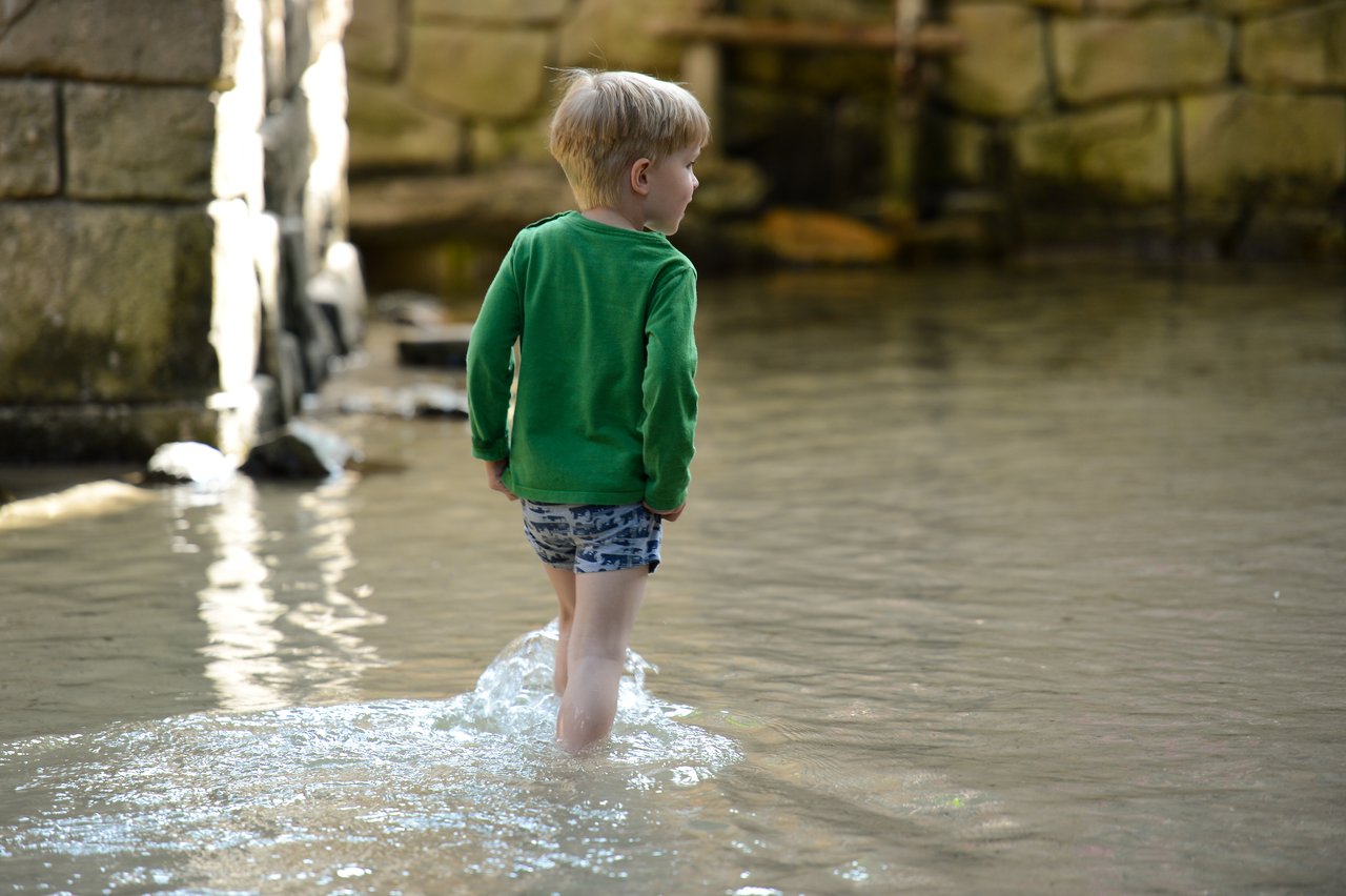 A young child in a green sweater walks through shallow water, creating small splashes.