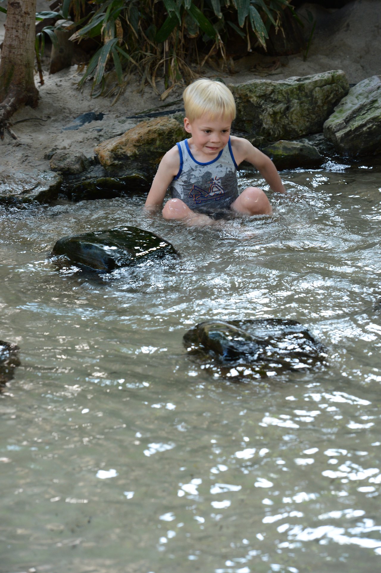 A young child sits in shallow water, surrounded by rocks, wearing a sleeveless shirt and looking focused.
