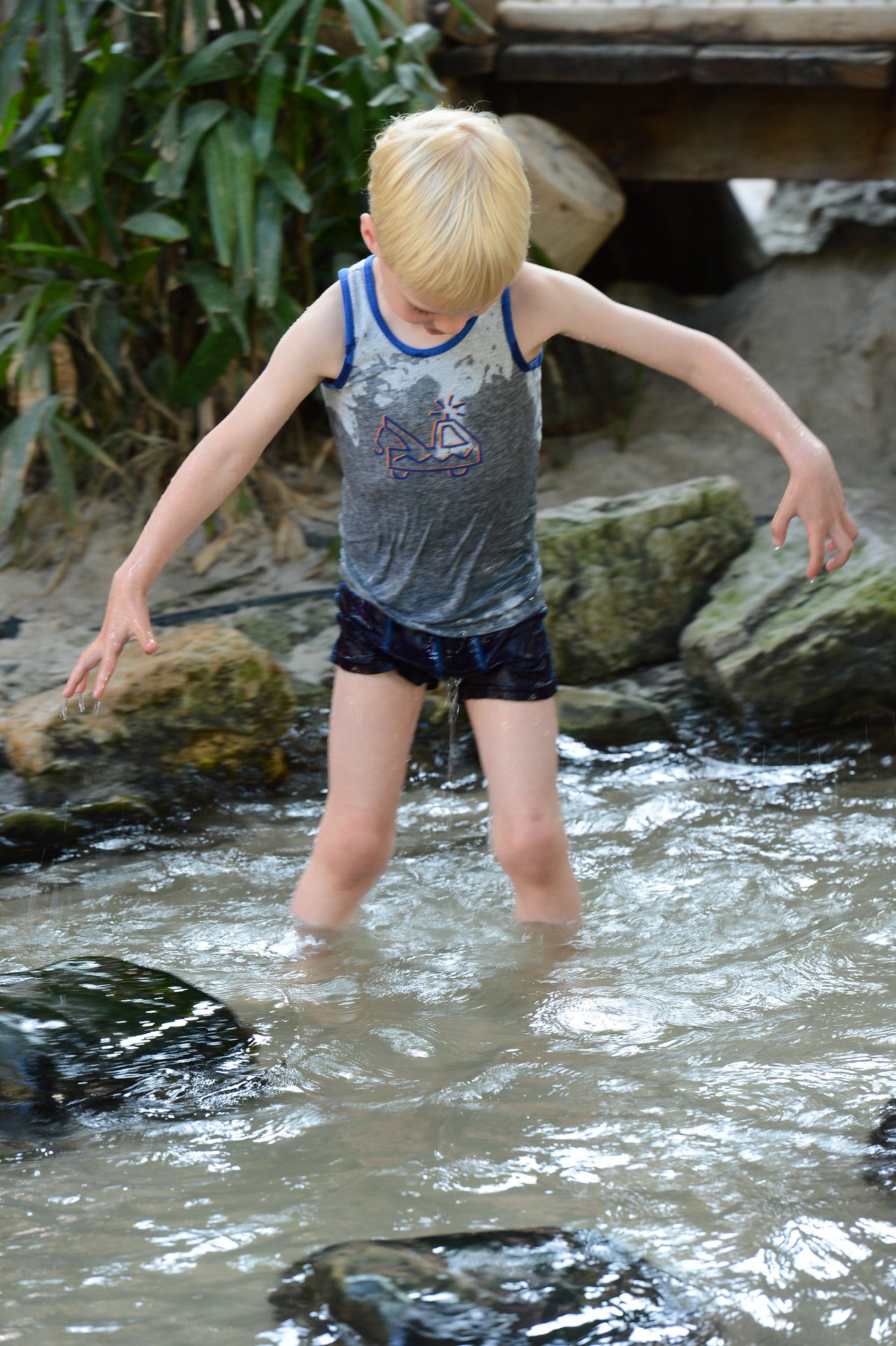 A child with wet clothes wades through shallow water, balancing carefully on uneven rocks.