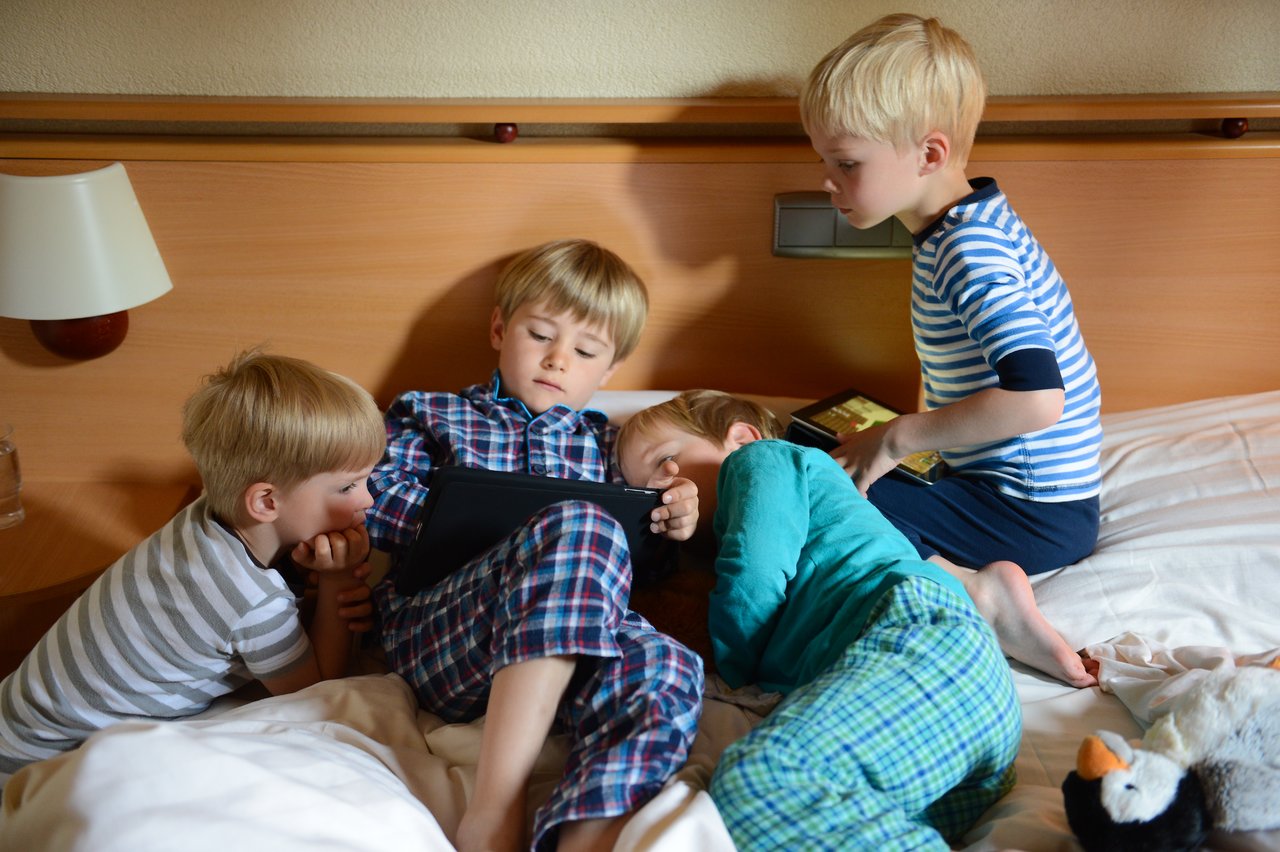 Four young children in pajamas sit and lie on a bed, focused on a tablet screen together.