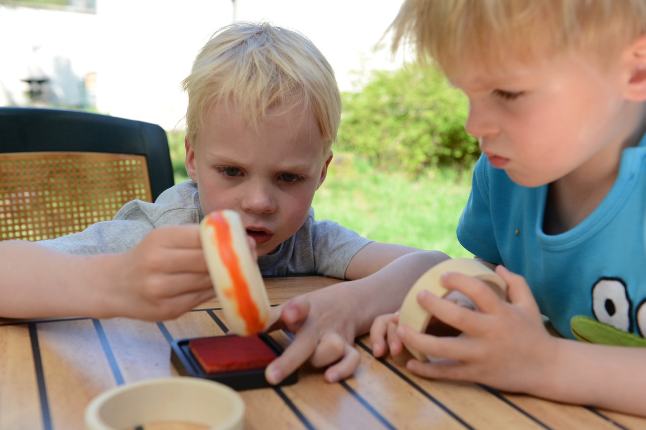 Two young children sit at a wooden table, stamping with ink and playing with toy food.