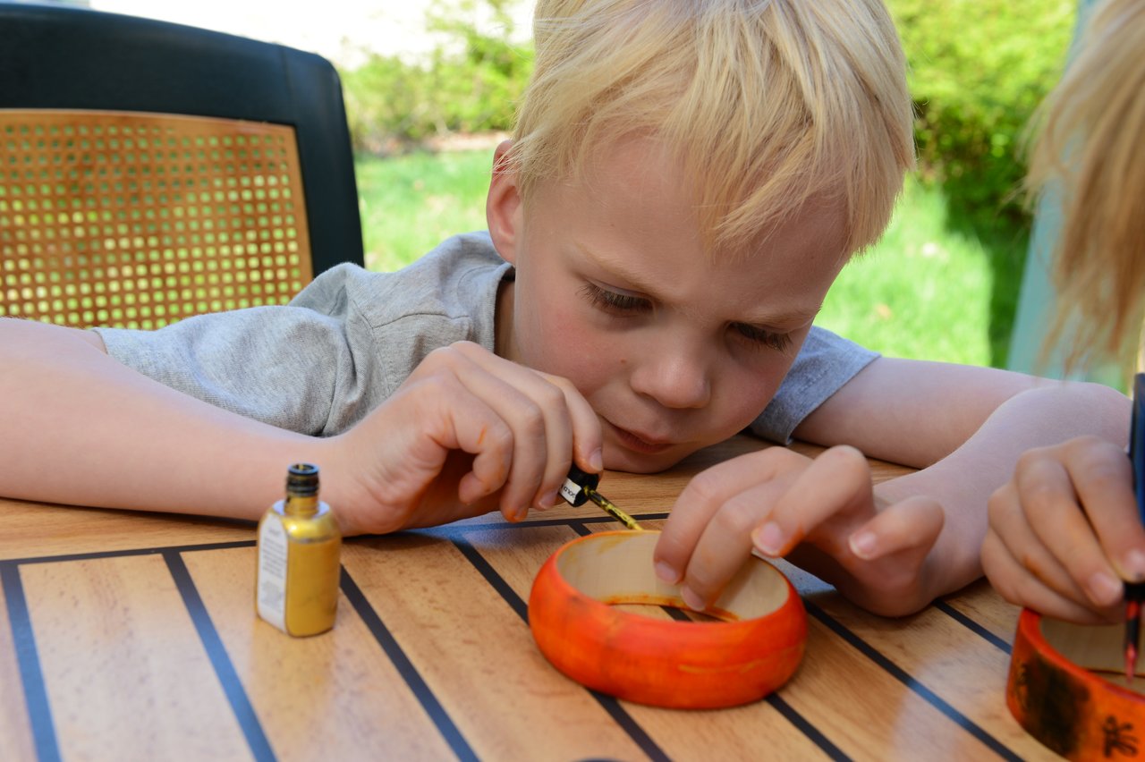 A young child carefully paints a wooden bracelet with a small brush at an outdoor table.