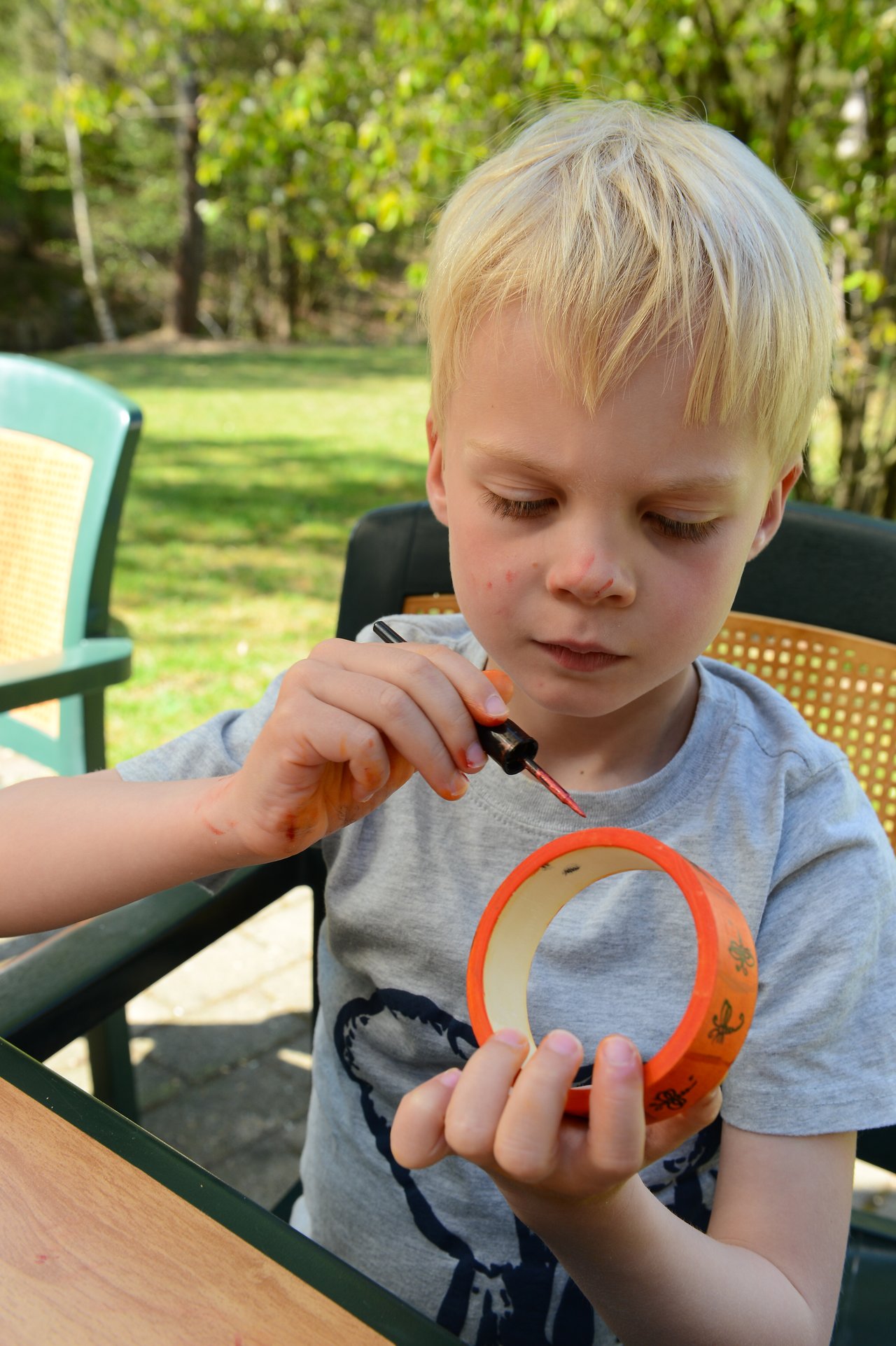 A young child carefully paints an orange tape roll while sitting at an outdoor table.