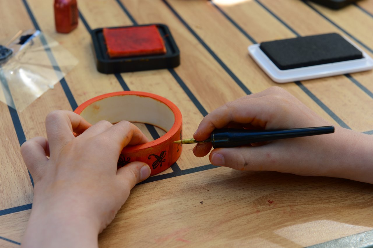 A child decorates a wooden bracelet with a black pen while sitting at a table with art supplies.
