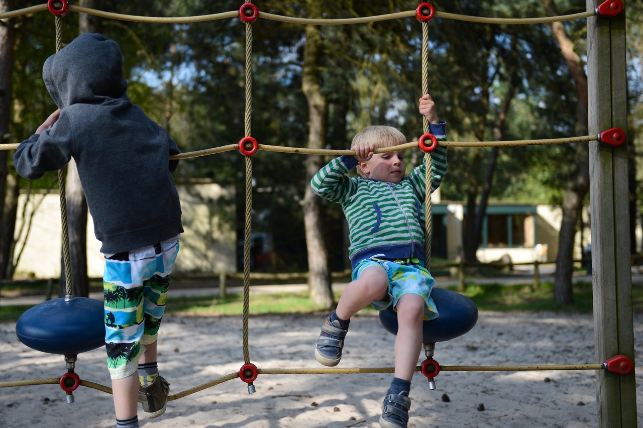 Two children climb a rope playground structure, holding onto the ropes and balancing on suspended seats.