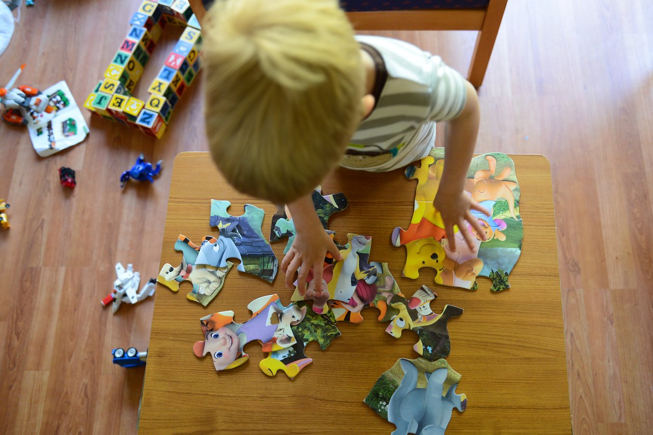 A child assembles a colorful puzzle with cartoon characters on a wooden table, surrounded by scattered toys.